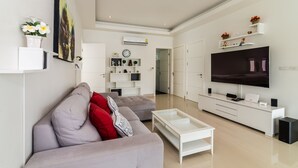 A modern living room with a grey sofa, a coffee table, and a flat-screen TV. There are white shelves with decorative items and a door leading to another room. - Orchid Paradise Homes OPV210 (Hua Hin)