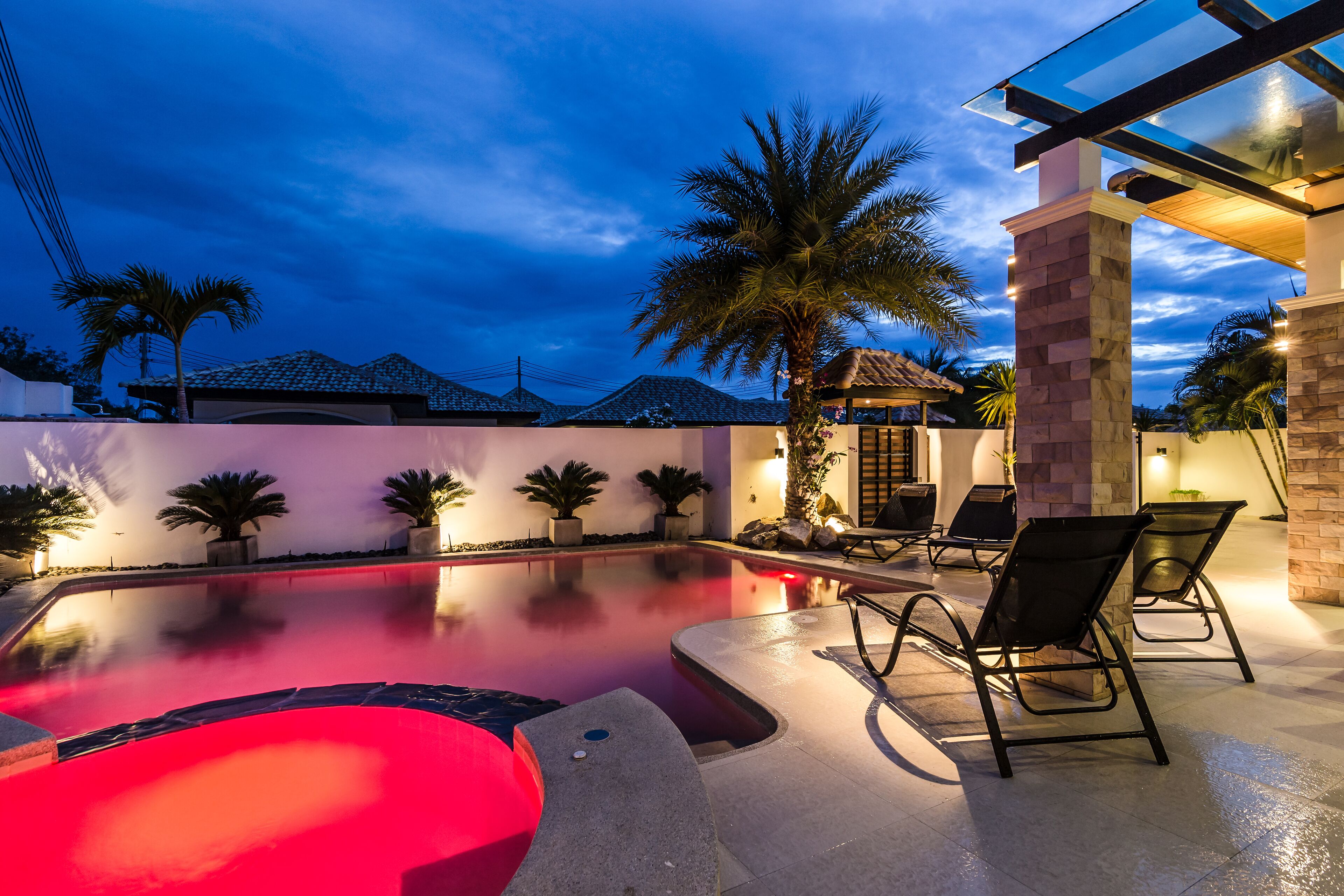 A swimming pool with a red-lit hot tub, surrounded by palm trees and lounge chairs.