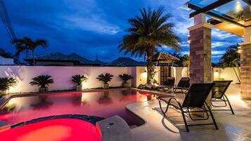 A swimming pool with a red-lit hot tub, surrounded by palm trees and lounge chairs.