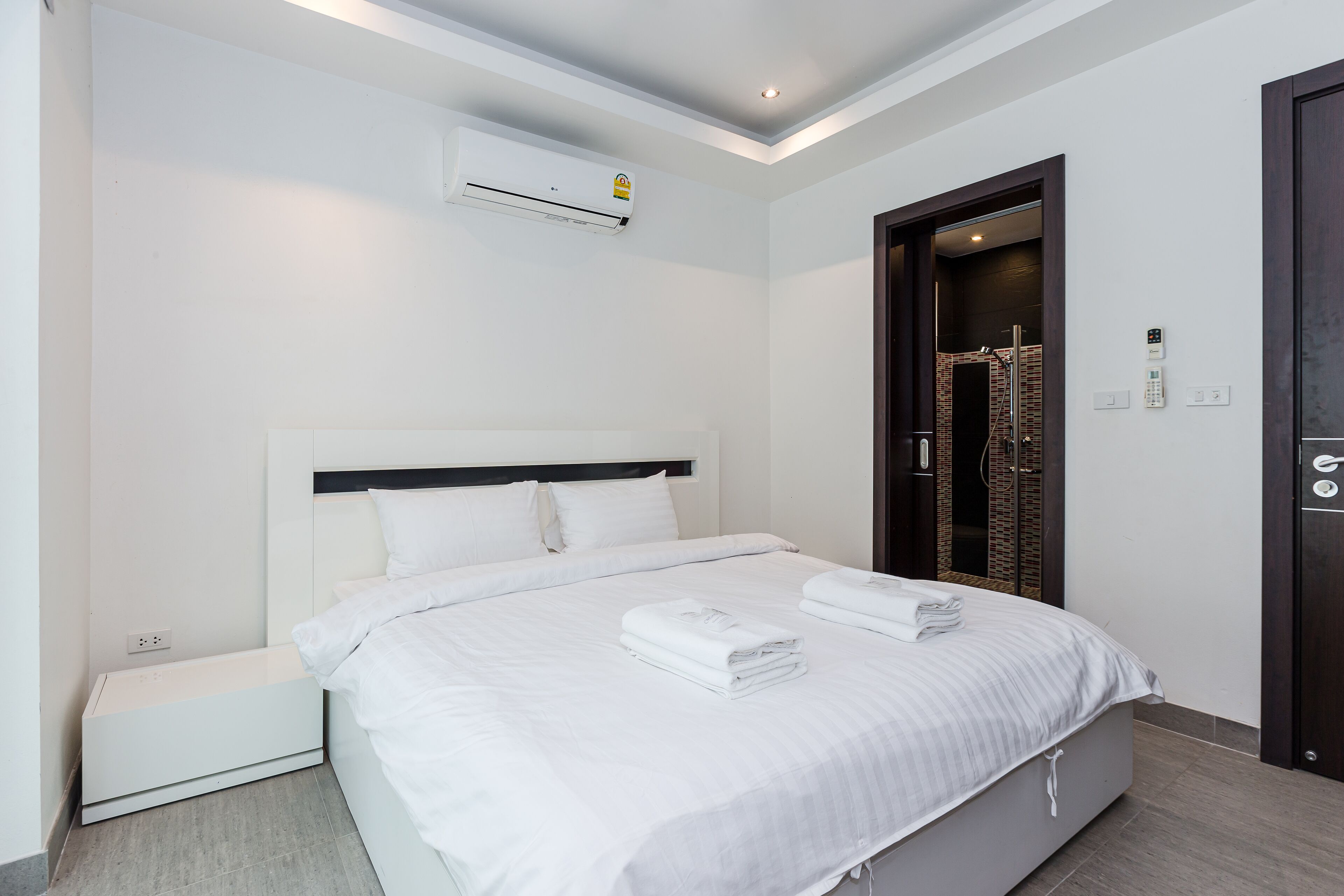 Een moderne hotelkamer met een groot bed, witte beddengoed, een airconditioningapparaat, een ingebouwde verwarming, een donkerhouten deur en een klein nachtkastje.