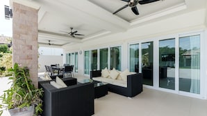 A covered outdoor patio with black wicker furniture, glass-top coffee table, ceiling fans, and large sliding glass doors. - Orchid Paradise Homes OPV219 (Hua Hin)