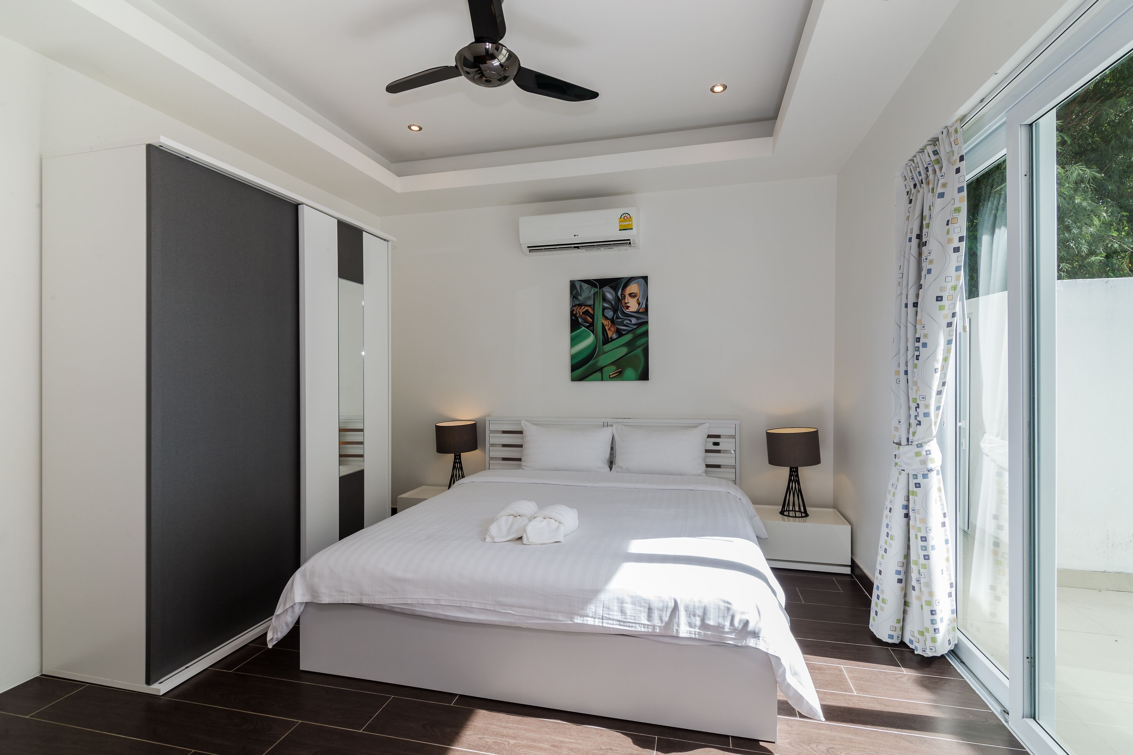 A modern bedroom with a large bed, a ceiling fan, an air conditioning unit, and a view of greenery outside.