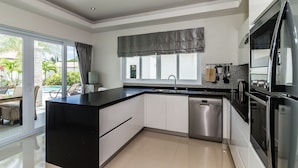 A modern kitchen with a black countertop, white cabinets, stainless steel appliances, and a large window overlooking a pool area. - Orchid Paradise Homes OPV220 (Hua Hin)