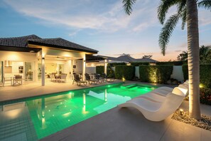 Un espace piscine extérieur moderne, doté d’un coin salon couvert, de chaises longues et d’une terrasse bien éclairée.