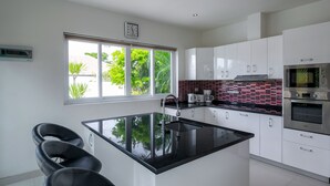 A modern kitchen with a central island, white cabinets, a red-tiled backsplash, and a window with a view of greenery.