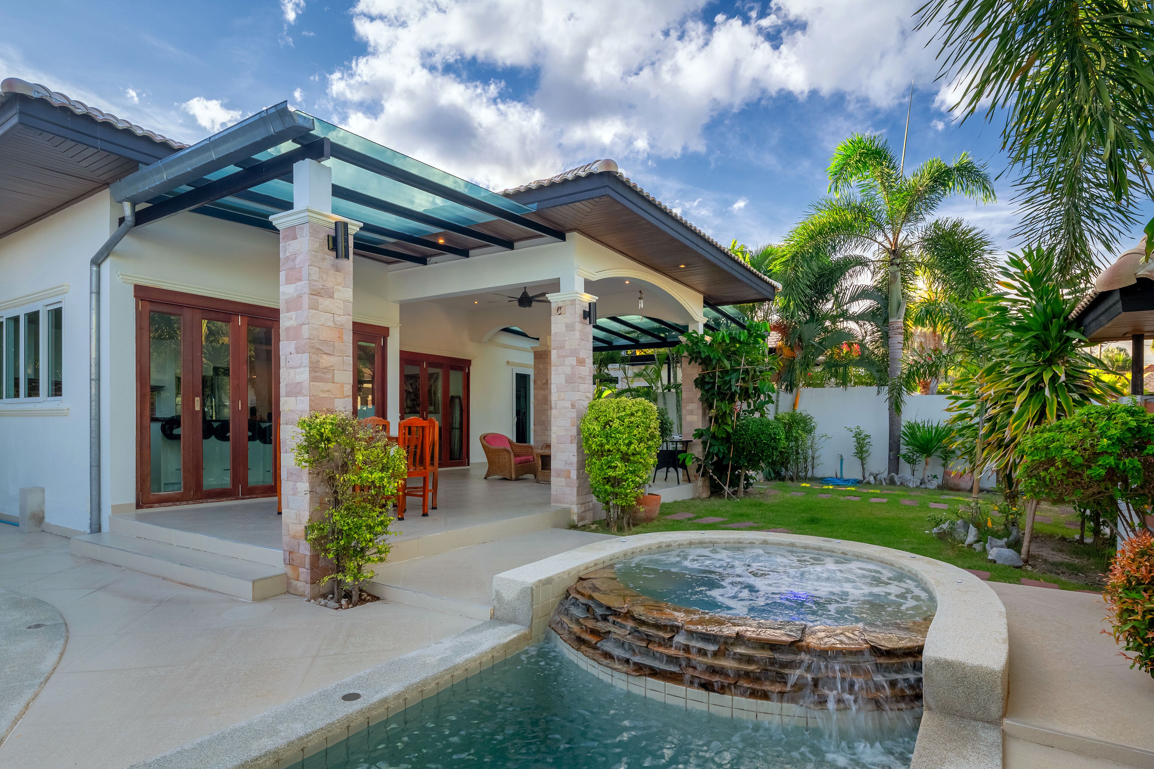 A modern house with a covered patio, a swimming pool with a waterfall feature, and lush tropical landscaping.