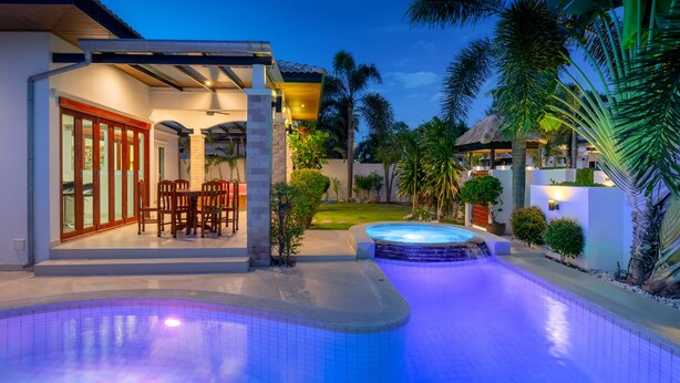 A swimming pool with a hot tub, a covered patio with chairs, and a well-maintained garden with palm trees.
