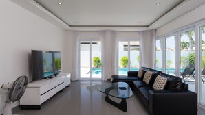A modern living room with a flat-screen TV, a glass coffee table, a black leather sofa, and a pool view through large windows. - Orchid Paradise Homes OPV315 (Hua Hin)