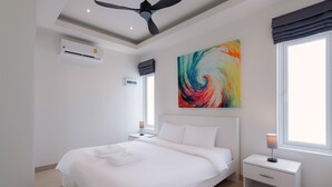 A modern bedroom with a large bed, a ceiling fan, an air conditioning unit, a bedside table with a lamp, and a colorful abstract painting on the wall. - Orchid Paradise Homes OPV515 (Hua Hin)