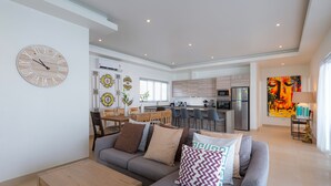 A modern living room with a sofa, coffee table, and decorative pillows. A kitchen with a dining area is visible in the background. - Orchid Paradise Homes OPV515 (Hua Hin)