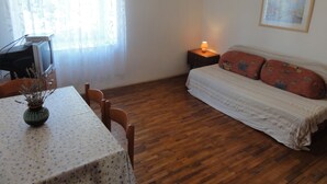 Una habitación con piso de madera, una cama con almohadones estampados, una mesa con mantel floreado, un florero con flores secas, una lámpara en una mesita de noche y un cuadro en la pared.