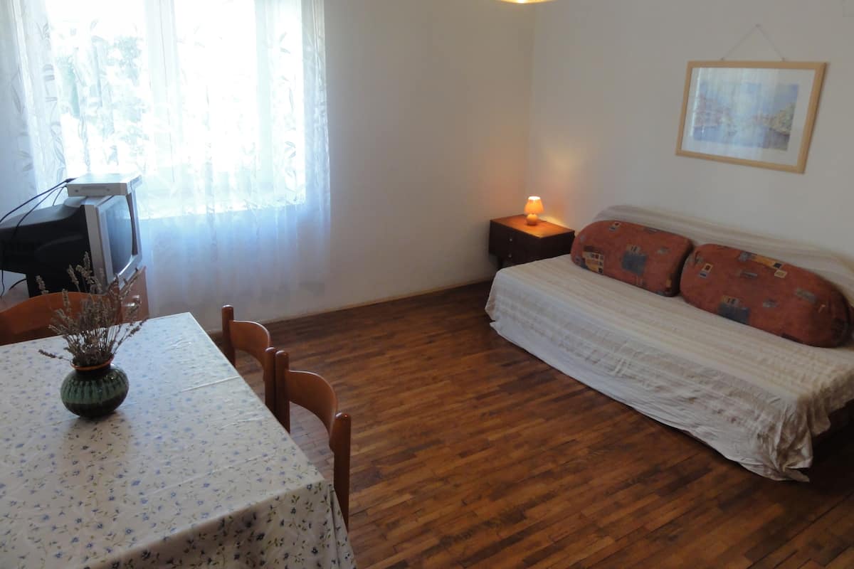 A room with a wooden floor, a bed with patterned pillows, a table with a floral tablecloth, a vase with dried flowers, a lamp on a bedside table, and a framed picture on the wall.