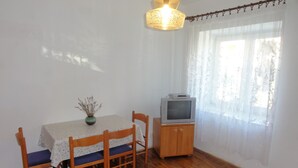 Un área de comedor con una mesa, sillas, un televisor sobre un mueble bajo y una ventana con cortinas ligeras.