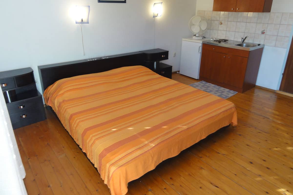 A bedroom with a bed, bedside tables, a nightstand, a small kitchenette with a sink and stove, and a bathroom with a toilet and sink.