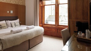 Ein Hotelzimmer mit einem hölzernen Kopfteil, einem Bett mit beiger Decke, einem Holztisch mit Kaffeemaschine und einem Fenster mit Vorhängen.
