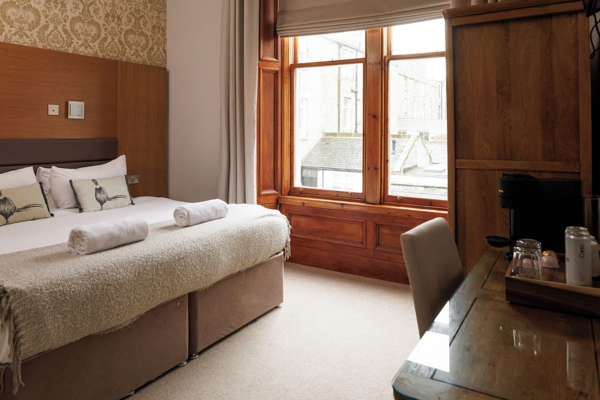Une chambre d’hôtel avec une tête de lit en bois, un lit recouvert d’une couverture beige, un bureau en bois avec une machine à café et une fenêtre avec des rideaux.