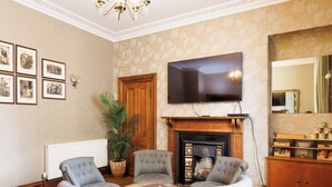 Ein Wohnzimmer mit zwei grauen Sesseln, einem Holztisch, einem Kamin, einem Flachbildfernseher, gerahmten Bildern an der Wand und einem großen Spiegel.