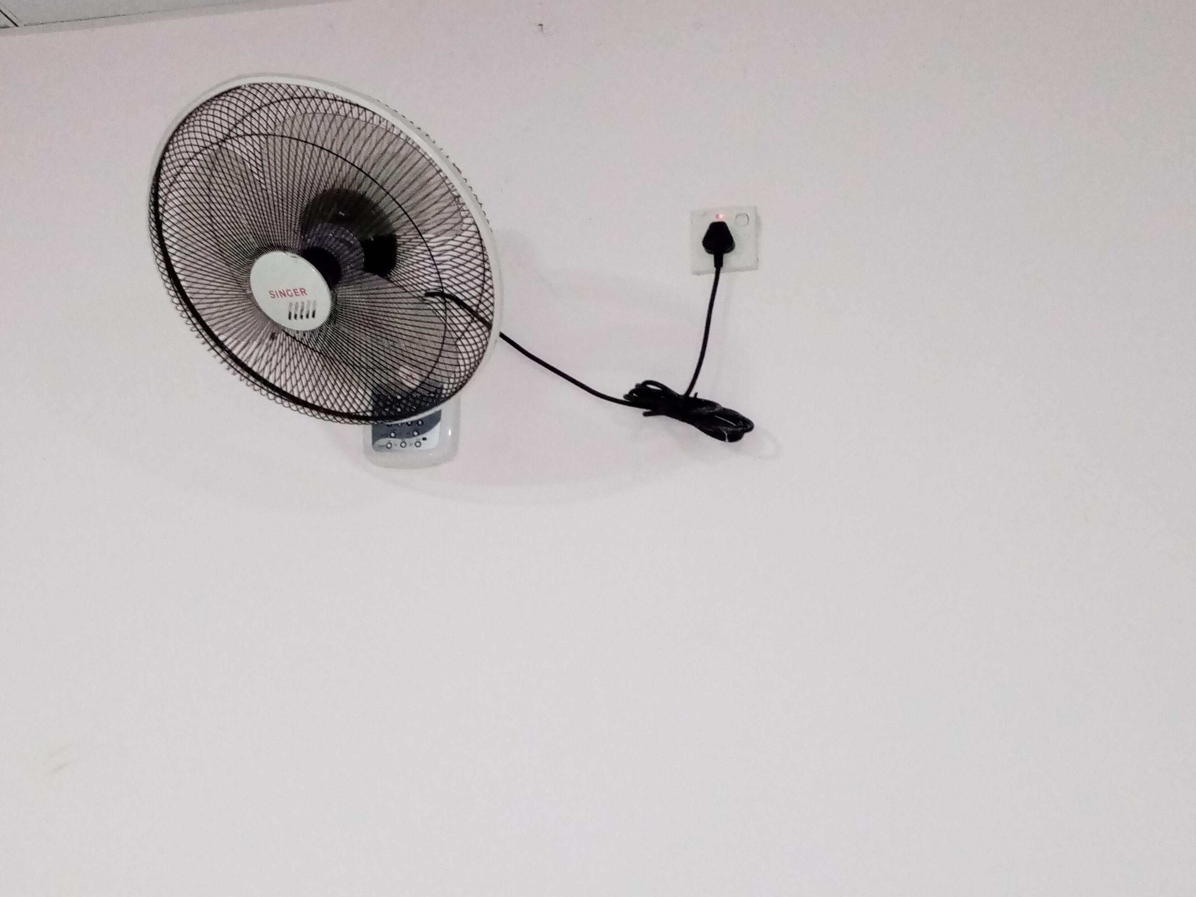 A standing fan with a power cord plugged into a wall socket.