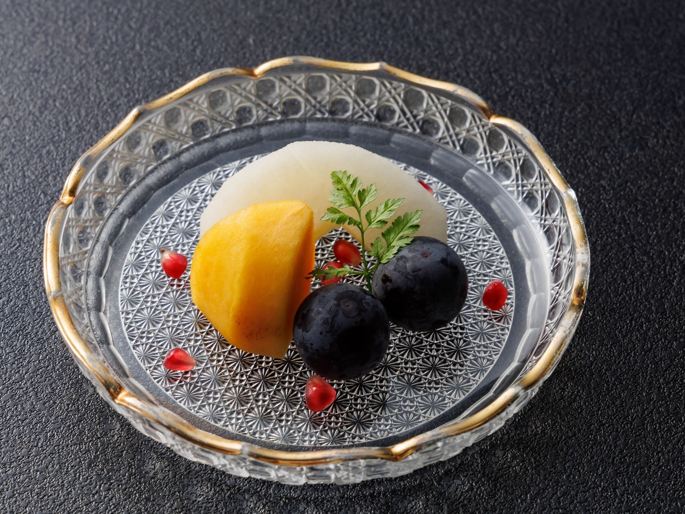 A clear glass plate with a geometric pattern, a yellow fruit, two dark round fruits, and a green sprig on a textured black surface.