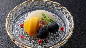 A clear glass plate with a geometric pattern, a yellow fruit, two dark round fruits, and a green sprig on a textured black surface.