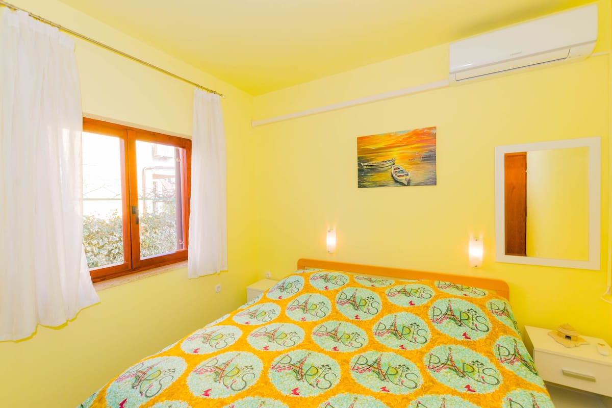 A room with a bed, a window with curtains, a painting on the wall, and a bedside table with a lamp.