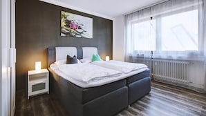 Quarto moderno com cabeceira escura, mesa de cabeceira branca e uma janela grande com cortinas leves.