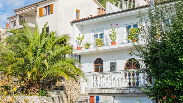 Un immeuble de deux étages avec une façade blanche, des balcons ornés de plantes en pot et un mur de pierre surmonté d’un palmier.