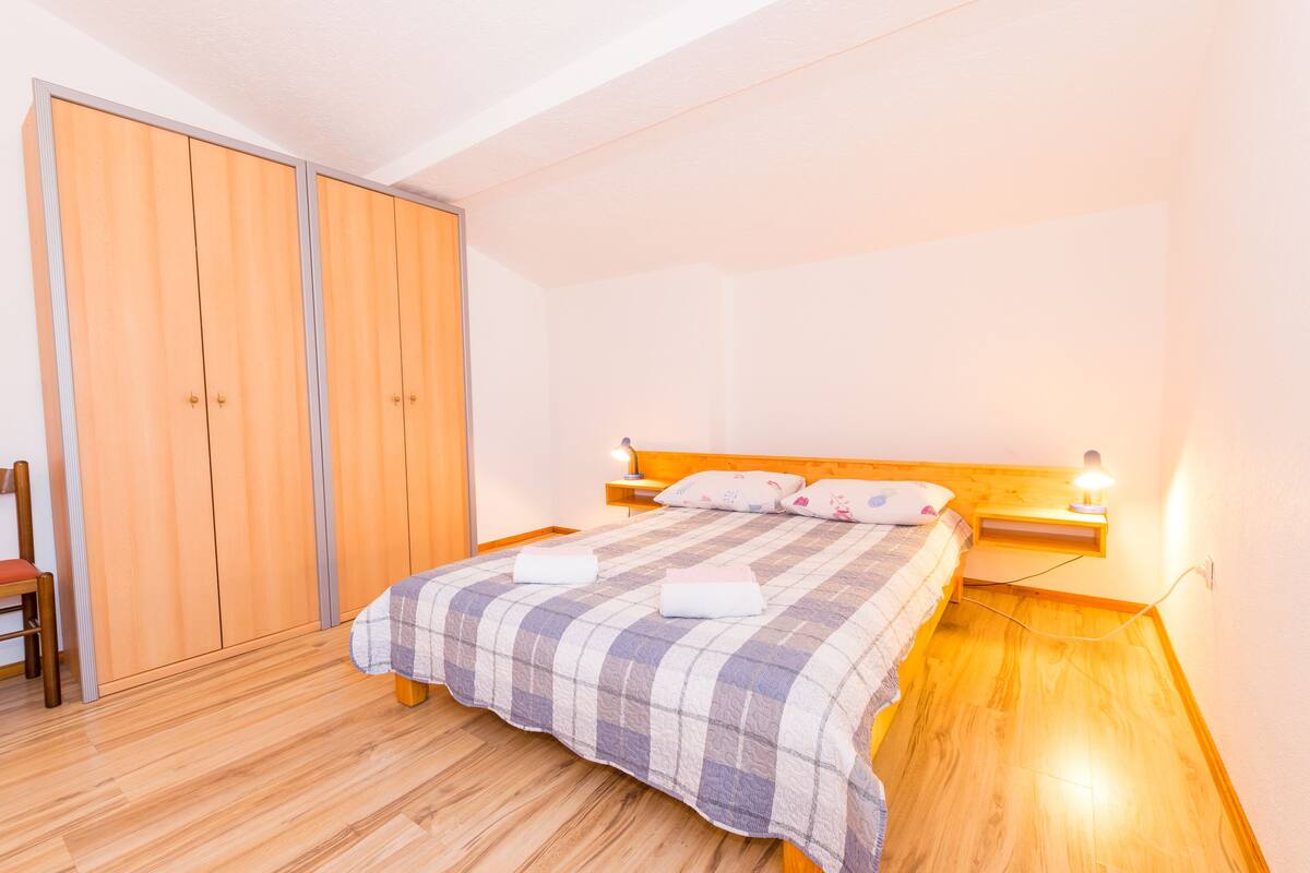 A bedroom with a wooden bed, a nightstand with a lamp, a wooden wardrobe, and a chair.