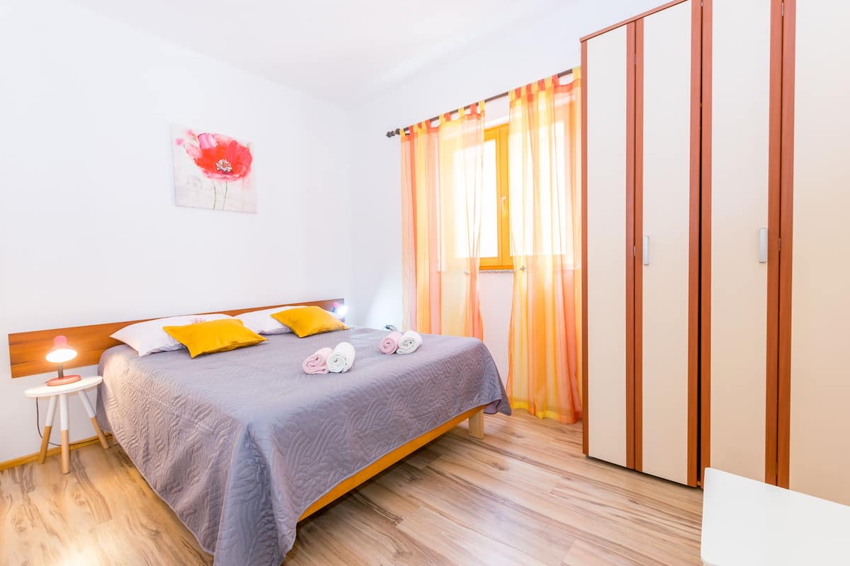 A bedroom with a bed, a nightstand, a wooden wardrobe, and a window with curtains.