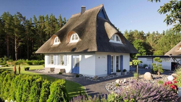 Une maison avec un toit de chaume, des murs blancs et une porte bleue. Elle est entourée d’un jardin bien entretenu, où poussent différentes plantes et fleurs.