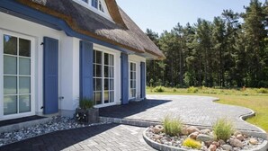 Une maison avec un toit de chaume, des volets bleus et une allée pavée menant à un espace aménagé avec des rochers et des plantes.