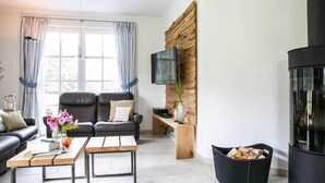 Un salon moderne avec un canapé en cuir noir, deux tables basses en bois, un téléviseur fixé à un mur en bois et une cheminée noire.