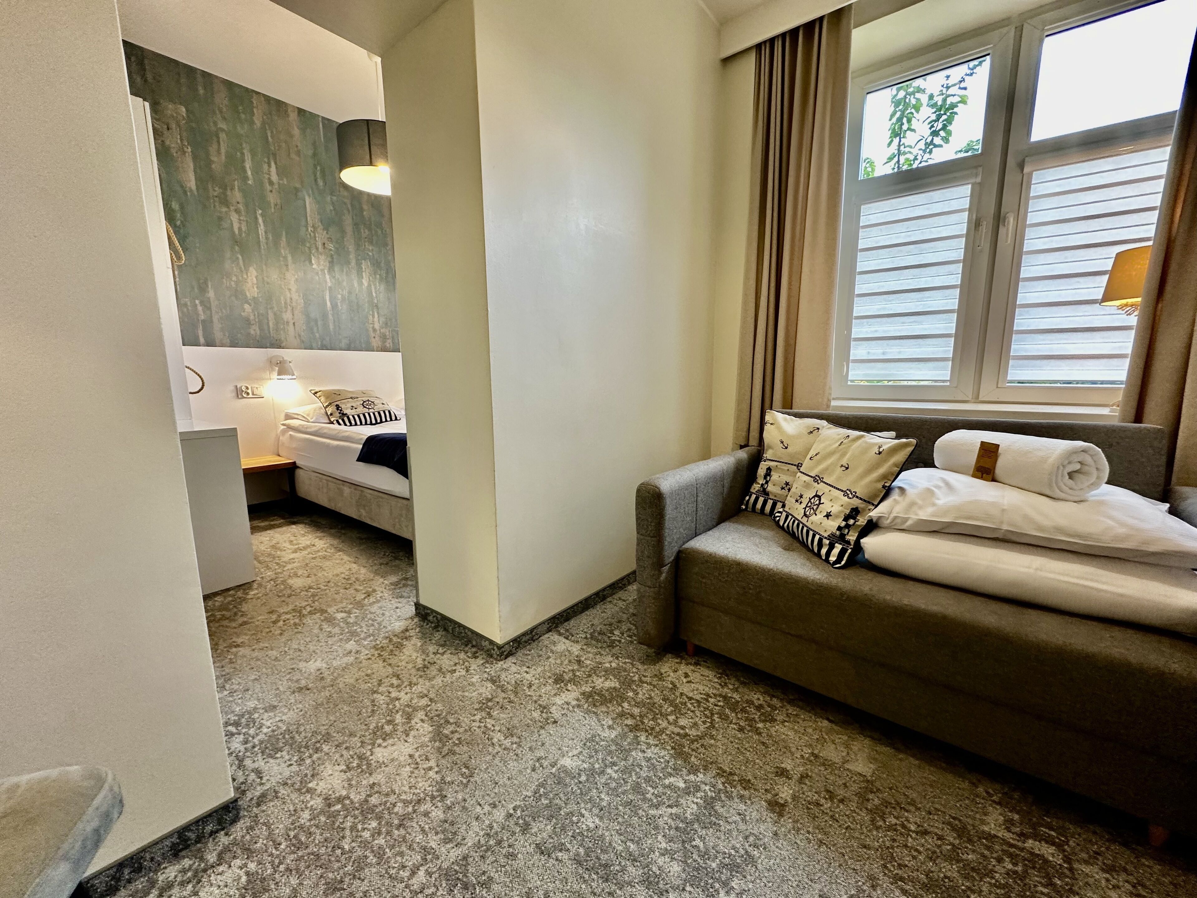 Une chambre d’hôtel comprenant un lit, un canapé, une fenêtre avec des rideaux, une lampe et un mur au décor texturé.