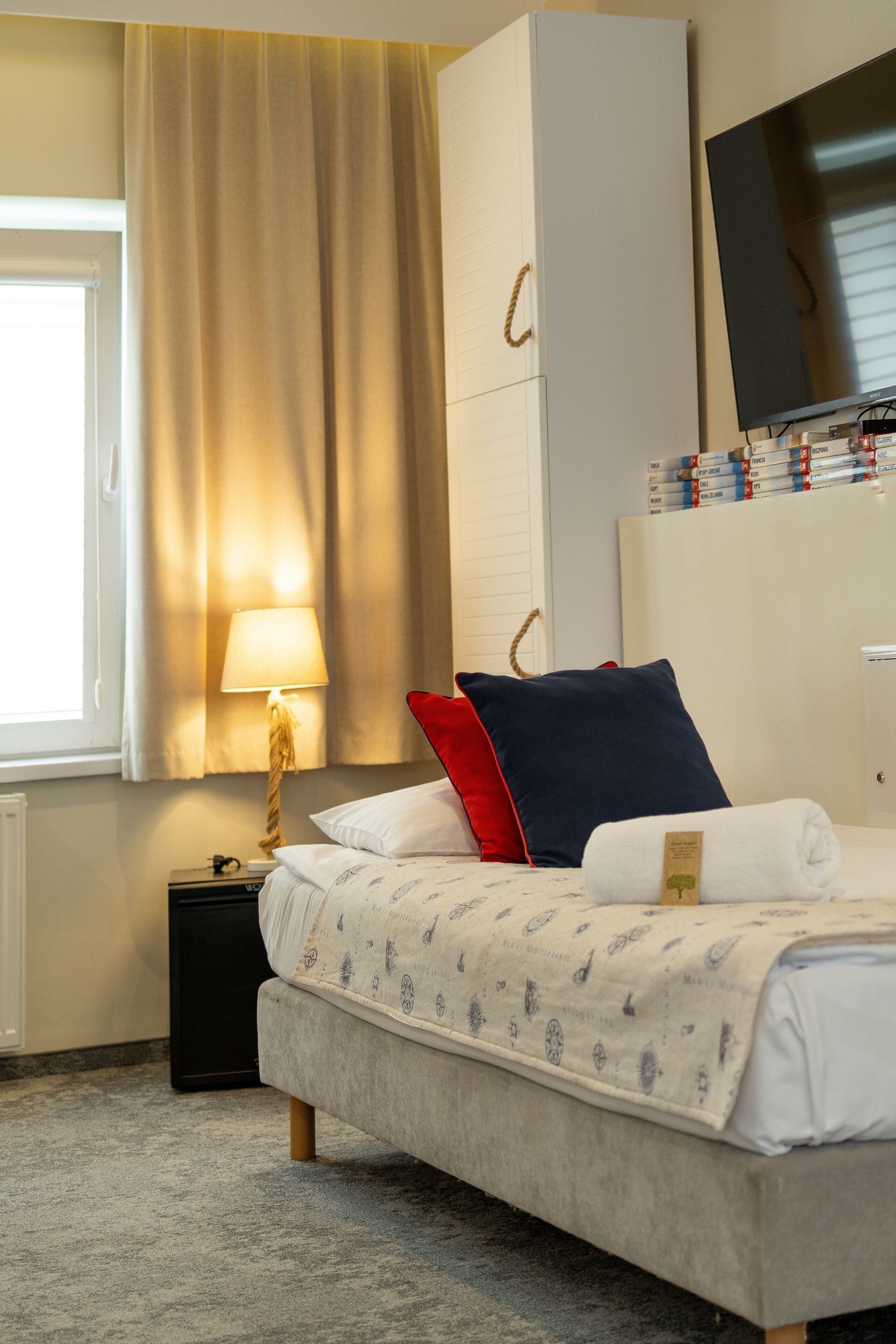 Una habitación de hotel ordenada con una cama, una mesita de noche con lámpara, un televisor y una estantería.