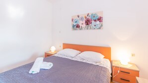 Una cama bien hecha con colcha azul y almohadas blancas, un cuadro floral en la pared y lámparas de noche sobre una mesita de madera.