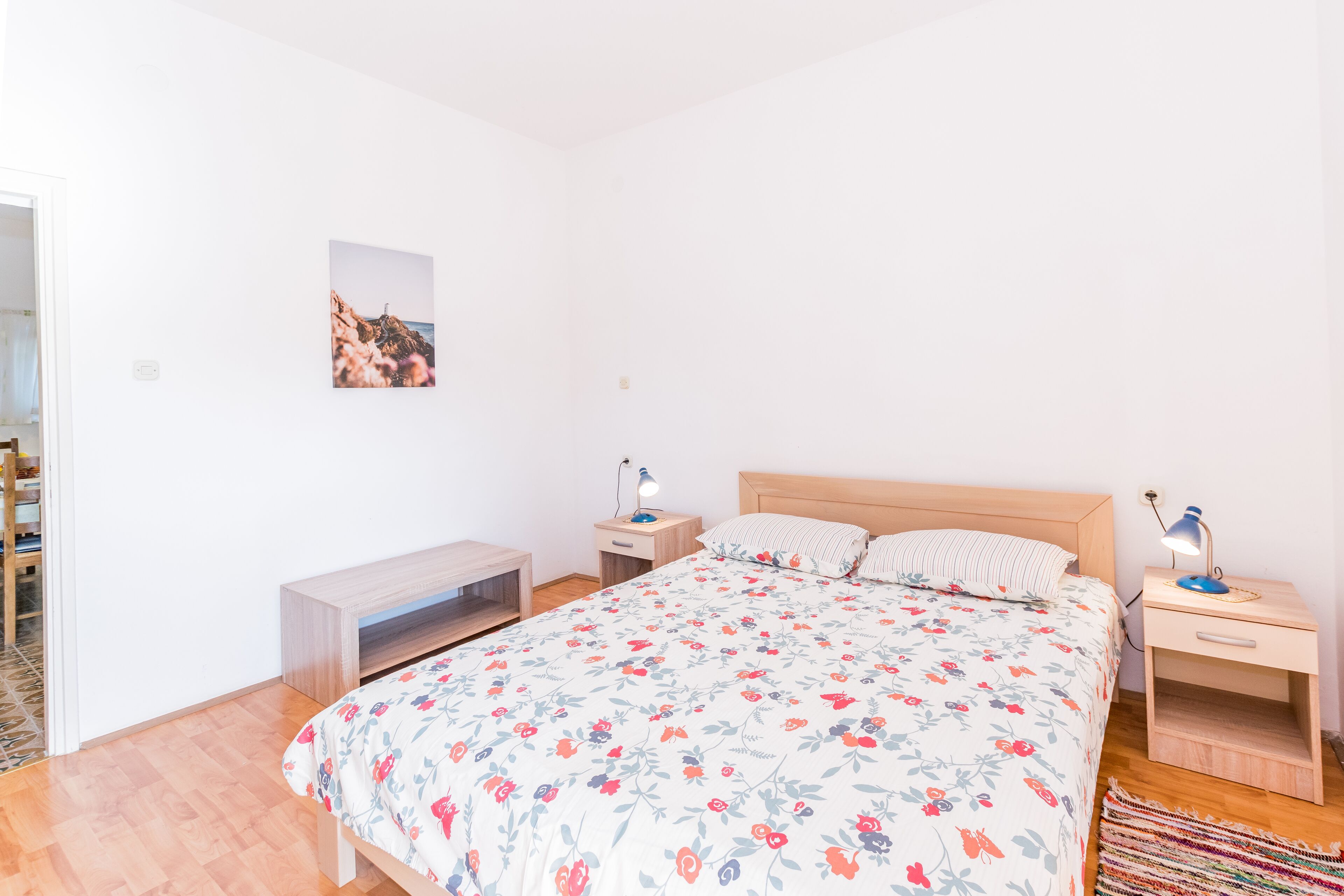 A bedroom with a floral bedspread, a wooden headboard, two bedside tables with lamps, a small bench, and a picture on the wall.