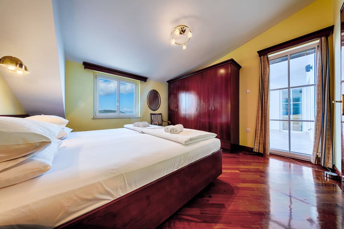 A large bed with white linens, a wooden wardrobe, a round mirror, and a view through a large window.