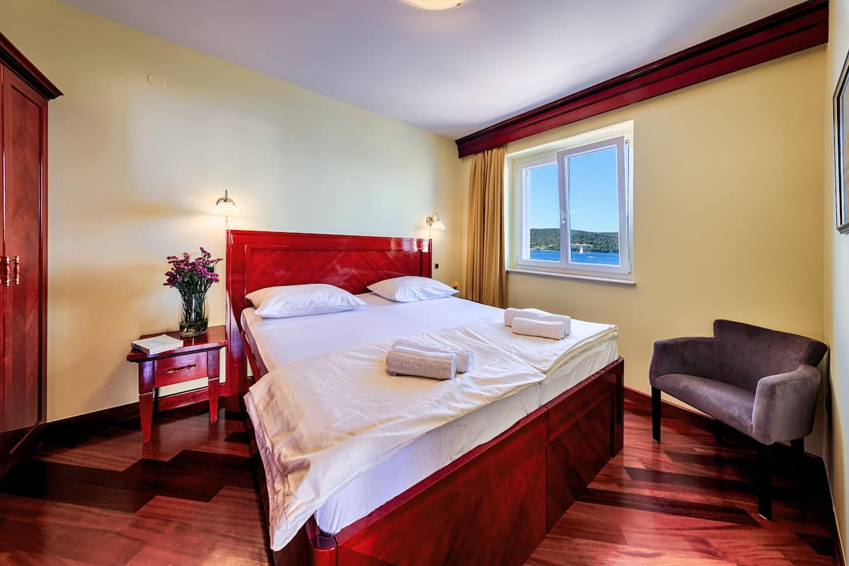 A hotel room with a wooden bed, a nightstand with a lamp, a chair, and a window with a view.