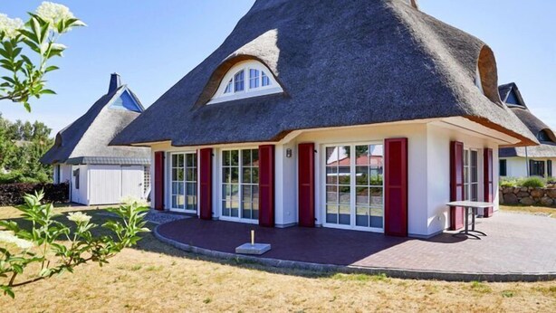 A traditional thatched-roof house with a white exterior and red shutters, featuring a large window and a small patio area.
