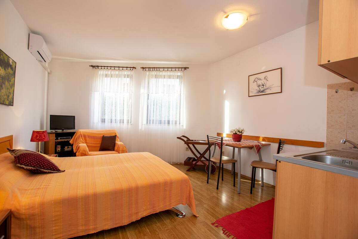 A compact hotel room with a bed, a desk, a chair, a small kitchen area, and a television.