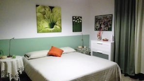 Ein ordentlich bezogenes Bett mit weißer Bettwäsche und einem orangefarbenen Kopfkissen, ein kleiner Nachttisch mit Lampe, eine Kommode und eine Wand mit einem großen Blumenbild und einem kleineren davon.