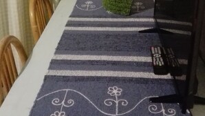 Un coin repas avec une table recouverte d’une nappe à rayures, un vase avec des fleurs, un tableau accroché au mur et un meuble sur lequel est posée une télévision, avec une télécommande à proximité.