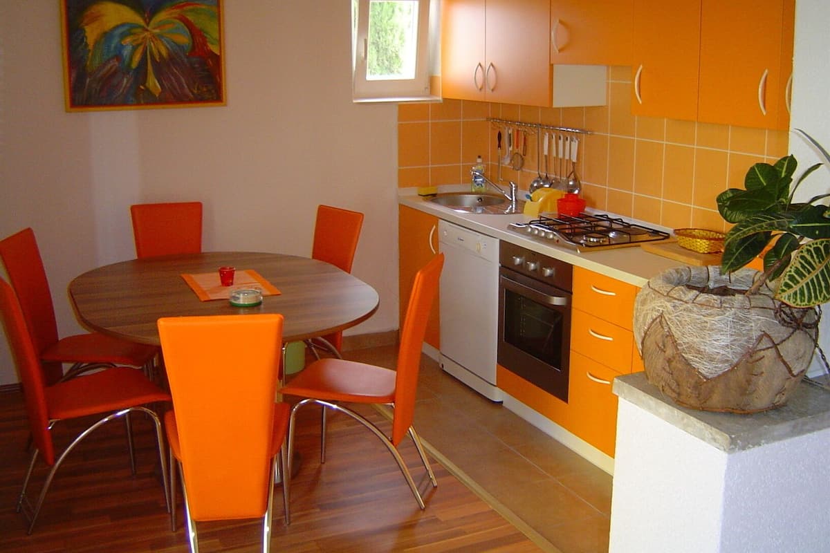 A compact kitchen with orange cabinets, a wooden dining table with orange chairs, a small window, and a painting on the wall.