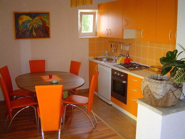 A compact kitchen with orange cabinets, a wooden dining table with orange chairs, a small window, and a painting on the wall.