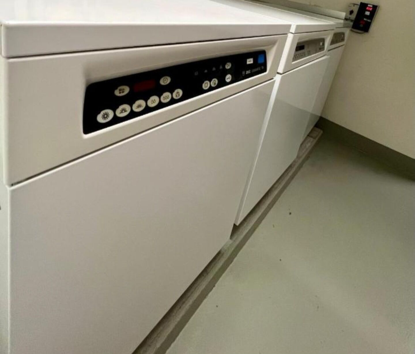 A white washing machine with a control panel, a wall-mounted control panel, and a tiled floor.