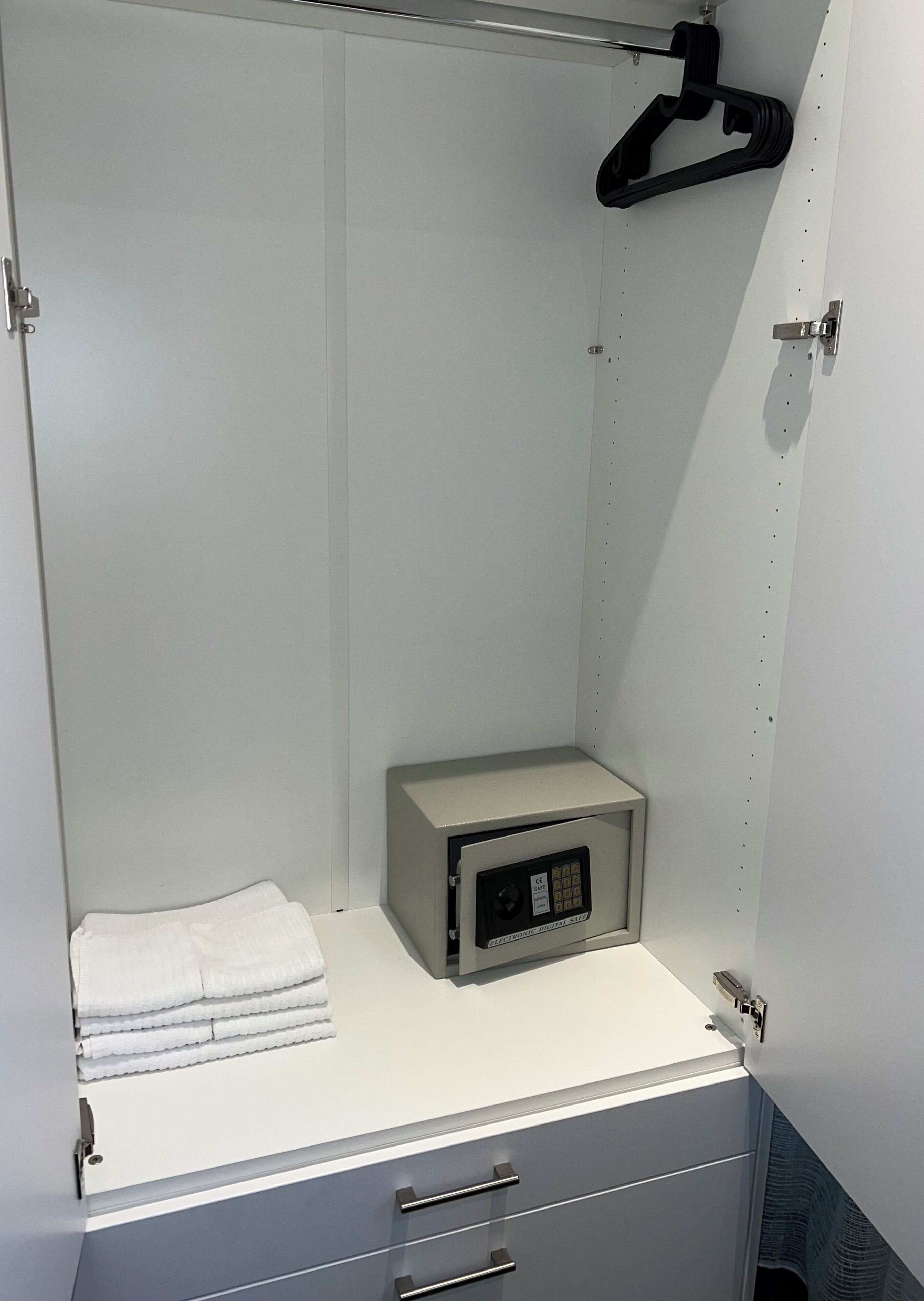 A small, enclosed room with a white cabinet containing a digital safe, a folded white towel, and a black hanger on the wall.
