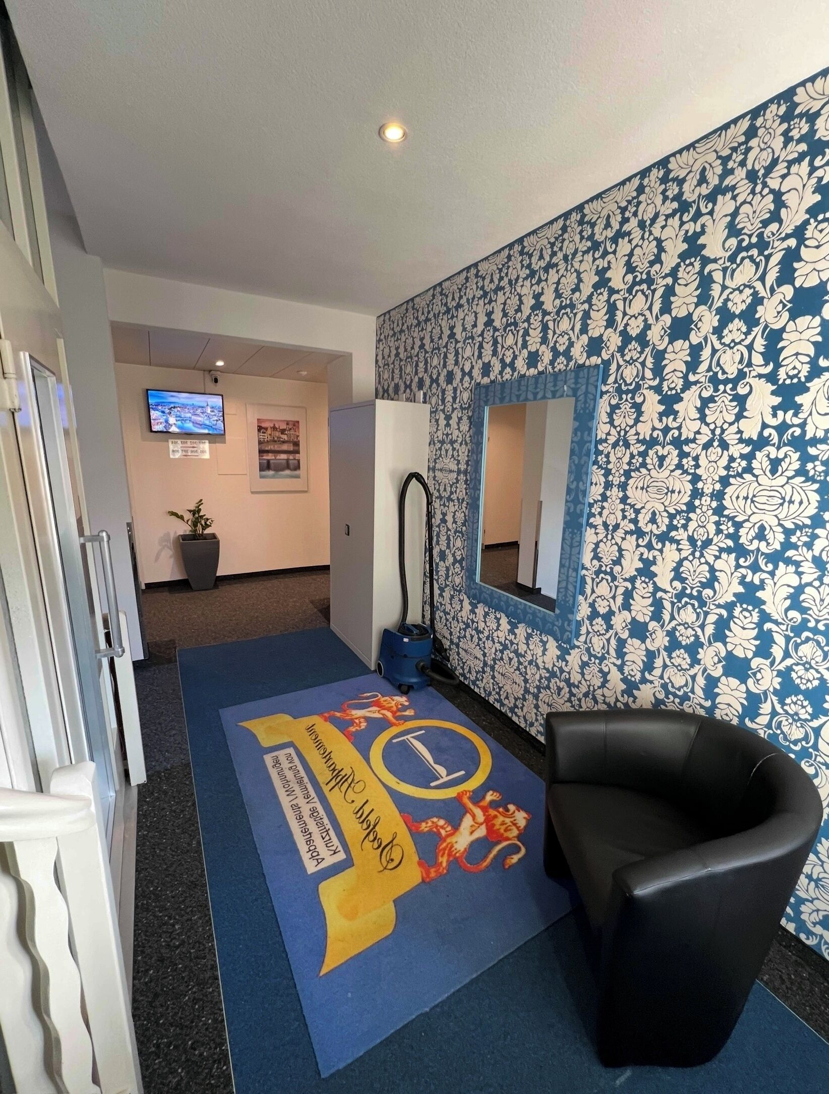 Ein Empfangsbereich mit einer Wand mit Muster, einem schwarzen Ledersessel, einem blauen Teppich mit Logo, einem Fernseher und einem Staubsauger.