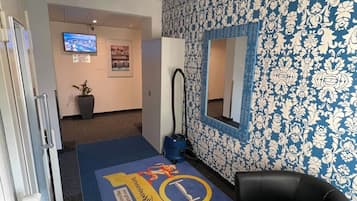 Ein Empfangsbereich mit einer Wand mit Muster, einem schwarzen Ledersessel, einem blauen Teppich mit Logo, einem Fernseher und einem Staubsauger.