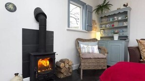 Un cuarto acogedor con un estufa de leña negra, un sillón de mimbre con cojín, un mueble azul con adornos, y una manta roja sobre el suelo.