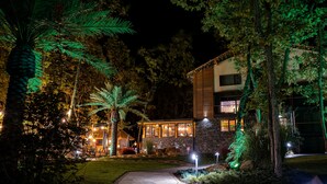 Una casa moderna con fachada de piedra, rodeada de exuberante vegetación e iluminada con luces colgantes.